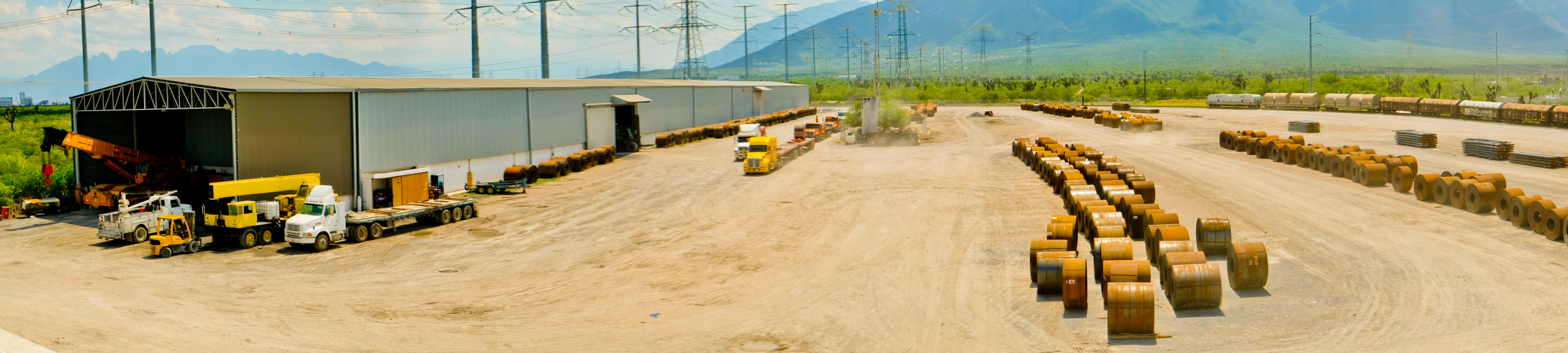 Terreno con bodega, vía ferroviaria, rollos de acero, maquinaria especializada, grúas hidráulicas y camiones de carga.